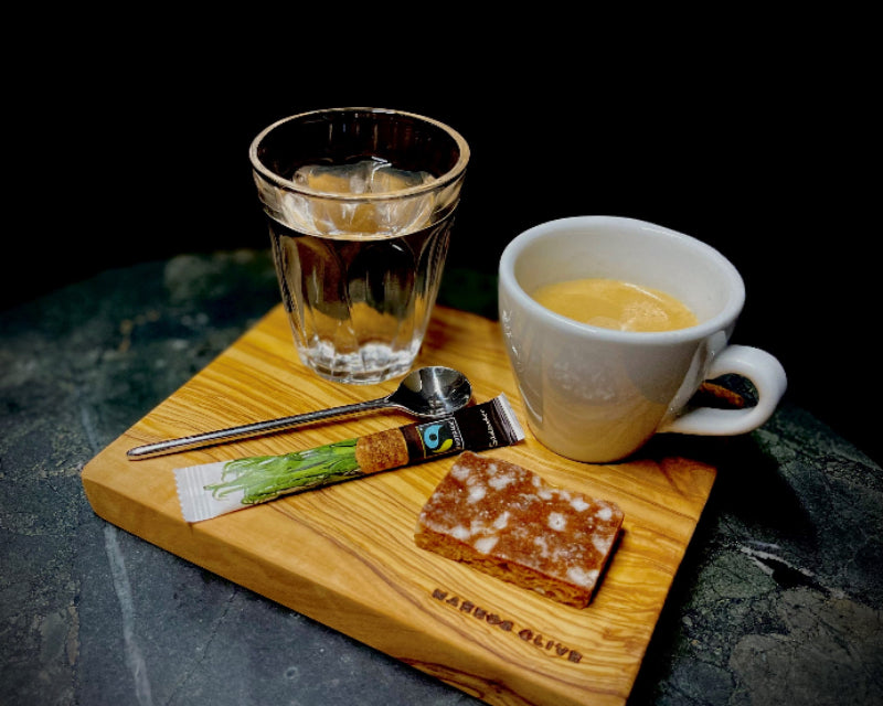 Ein elegantes Espressoset auf einem Tablett aus edlem Olivenholz mit dem Branding „MARKOS OLIVE“: ein Glas Wasser, eine Tasse Espresso, ein Fairtrade-Zuckerstick, ein kleiner Löffel und ein Stück Gebäck sind sorgfältig arrangiert – mediterraner Genuss mit Liebe zum Detail.