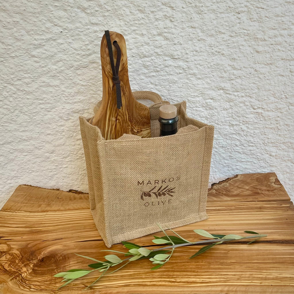Jute-Geschenktasche von MARKOS OLIVE gefüllt mit einer Flasche Ariston-Olivenöl, einem Glas Kalamata-Oliven und einem Schneidebrett aus Olivenholz