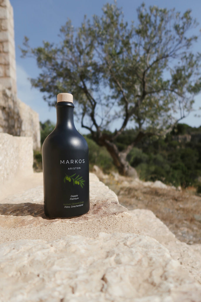 Schwarze MARKOS OLIVE Ariston Olivenölflasche auf Naturstein vor Olivenbaum in Griechenland bei Sonne