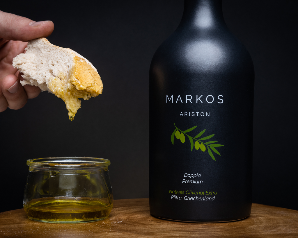 Flasche MARKOS ARISTON Premium Olivenöl 2024, abgefüllt in hochwertiger Frisches Brot wird in MARKOS OLIVE Ariston Olivenöl getaucht – edle schwarze Flasche mit griechischem Olivenzweig, typisch für die griechische Tradition.