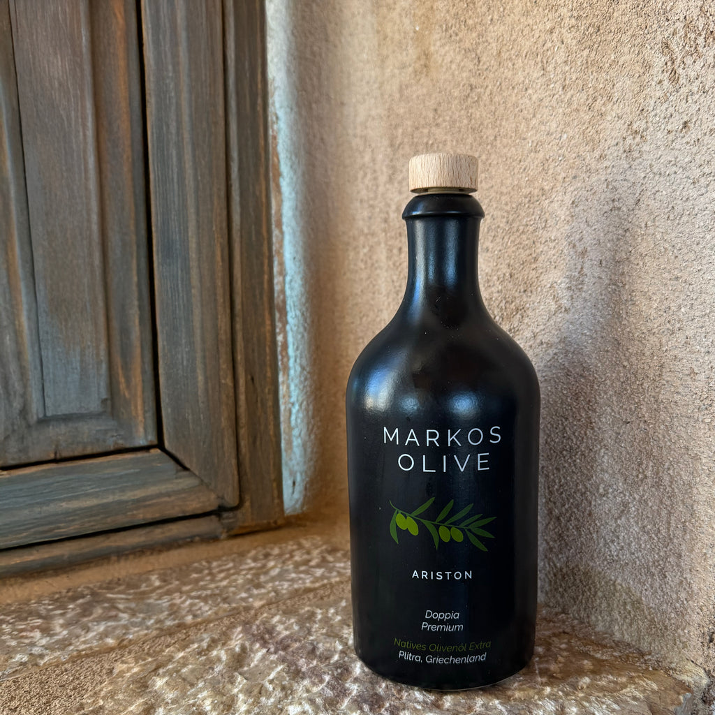 Schwarze Flasche MARKOS OLIVE Ariston Olivenöl auf Natursteinfensterbank neben Holzrahmen – hochwertiges Olivenöl aus Griechenland