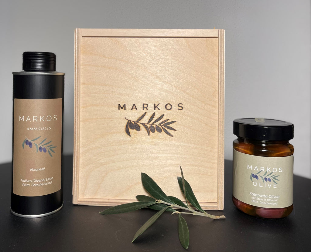 Geschenkbox von MARKOS OLIVE bestehend aus einer edlen Holzkiste, einer Flasche griechischem nativen Olivenöl extra (Koroneiki) und einem Glas Kalamata-Oliven mit Stein in Olivenöl. Vor der Box liegen Olivenzweige zur Dekoration.