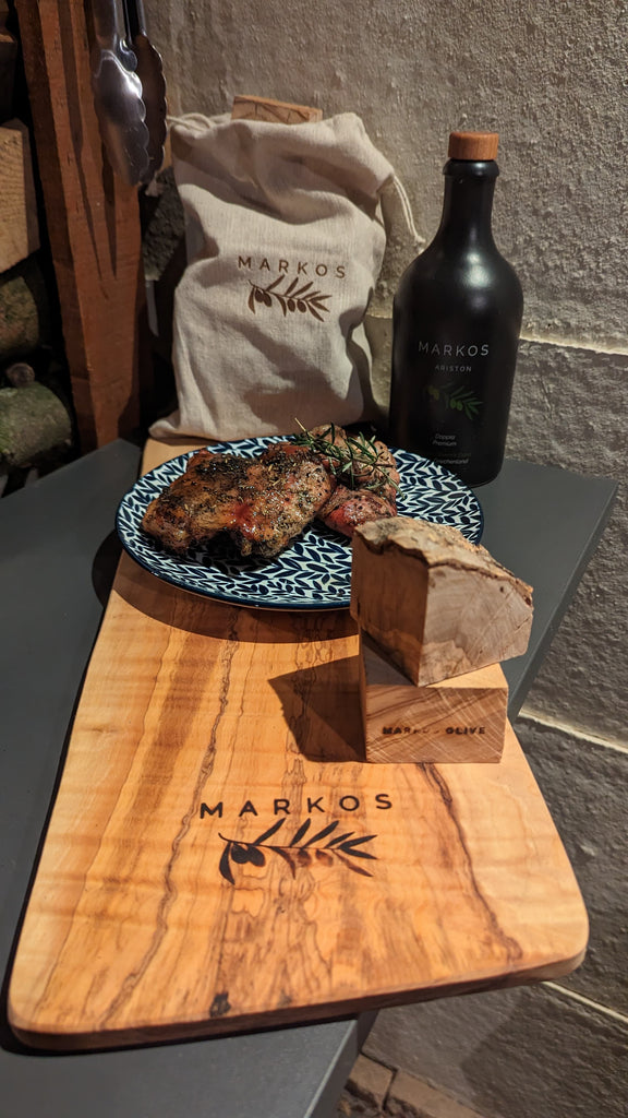 Gegrilltes Fleisch auf Olivenholzbrett mit MARKOS OLIVE Smoking Chunks, Olivenölflasche und Leinenbeutel – mediterranes BBQ-Setup
