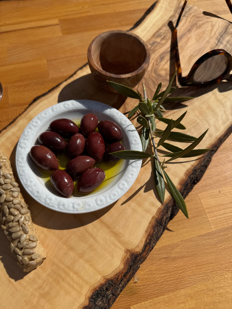 Stilvoll arrangierte mediterrane Szene mit einem Glas MARKOS Kalamata-Oliven in Olivenöl, begleitet von einem frischen Olivenzweig und natürlichem Lichtspiel. Die minimalistische Präsentation auf dunklem Untergrund unterstreicht die Authentizität und Qualität der griechischen Feinkost.