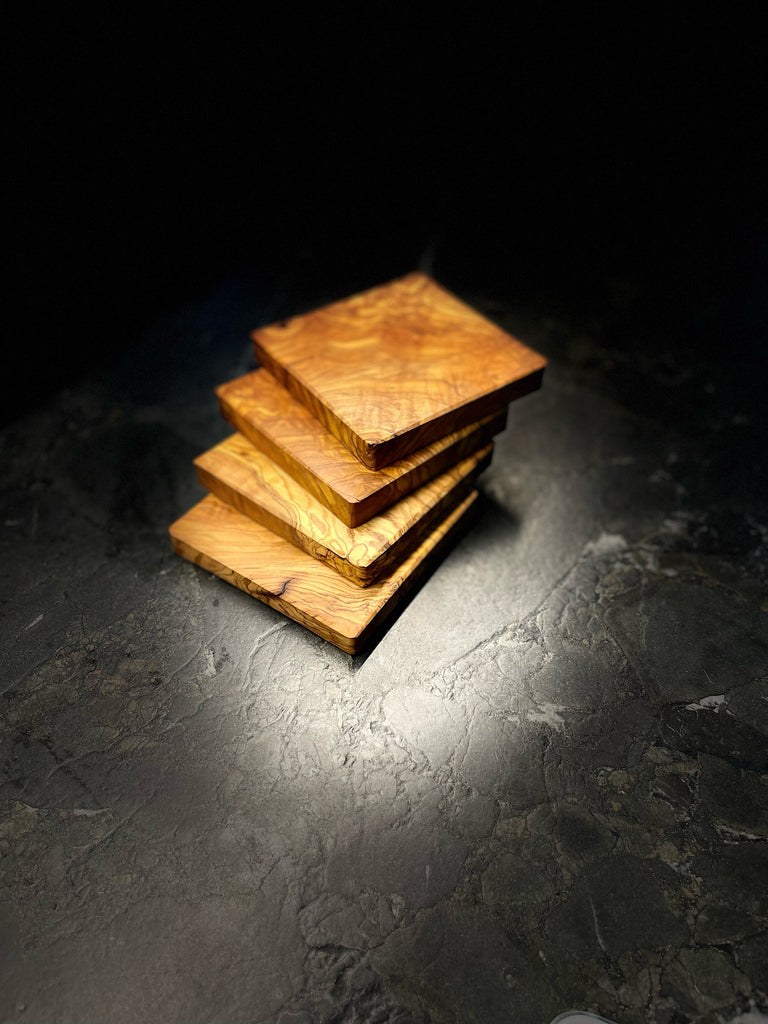 Vier quadratische Untersetzer aus edlem Olivenholz, jede mit einzigartiger Maserung und warmem Farbton. Auf dunklem Steinuntergrund präsentiert, bringen sie mediterranen Charme und handwerkliche Qualität auf jeden Tisch. Ideal für Gläser, Tassen oder kleine Servierideen.
