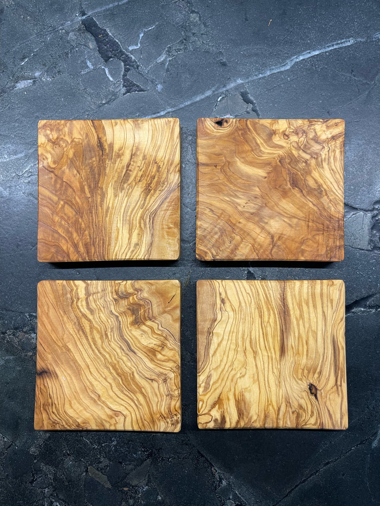 Vier quadratische Untersetzer aus edlem Olivenholz, jede mit einzigartiger Maserung und warmem Farbton. Auf dunklem Steinuntergrund präsentiert, bringen sie mediterranen Charme und handwerkliche Qualität auf jeden Tisch. Ideal für Gläser, Tassen oder kleine Servierideen.