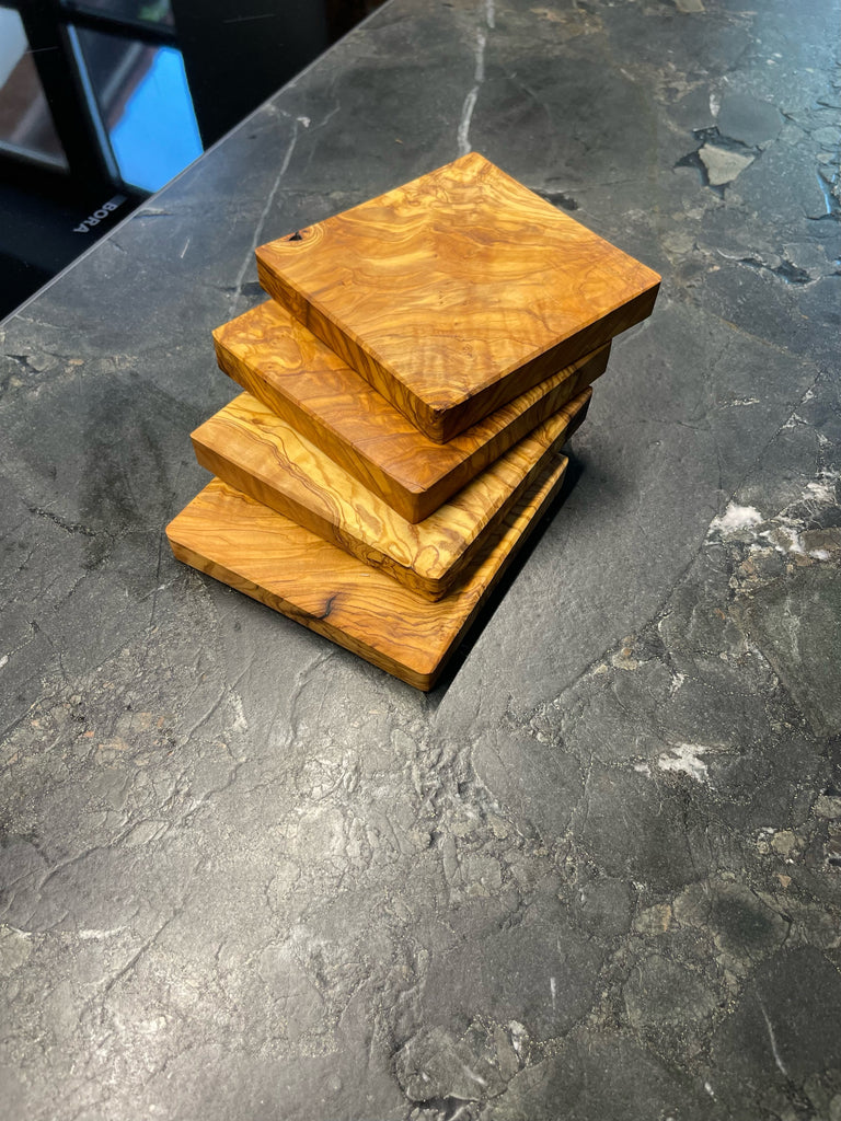 Vier quadratische Untersetzer aus edlem Olivenholz, jede mit einzigartiger Maserung und warmem Farbton. Auf dunklem Steinuntergrund präsentiert, bringen sie mediterranen Charme und handwerkliche Qualität auf jeden Tisch. Ideal für Gläser, Tassen oder kleine Servierideen.