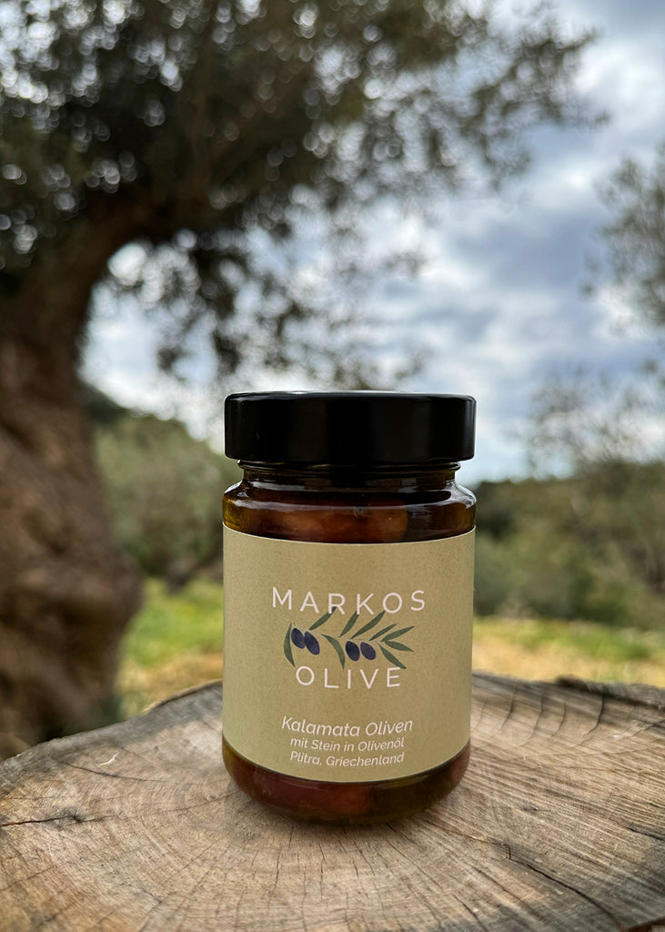 Ein Glas MARKOS Kalamata-Oliven mit Stein in Olivenöl steht auf einem alten Baumstumpf, im Hintergrund ein unscharfer Olivenbaum und griechische Landschaft. Das Etikett zeigt Herkunft und Natürlichkeit: Plitra, Griechenland. Authentische Präsentation mitten im Olivenhain.