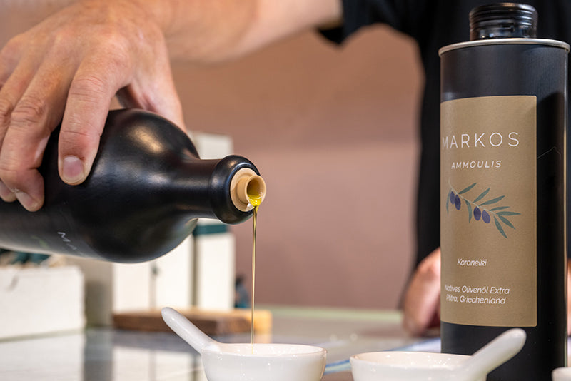 Eine Hand gießt hochwertiges MARKOS AMMOULIS Olivenöl aus einer schwarzen Flasche in ein kleines weißes Verkostungsschälchen. Daneben steht eine Dose mit dem Etikett „Natives Olivenöl Extra – Koroneiki, Plitra, Griechenland“. Szene aus einer professionellen Verkostungssituation.