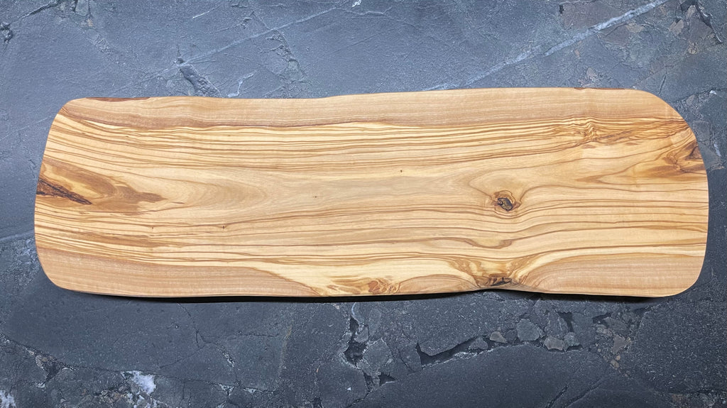 Massives Schneidebrett aus Olivenholz mit sanft geschwungener Naturform und ausdrucksstarker Maserung, auf dunklem Steinuntergrund präsentiert.