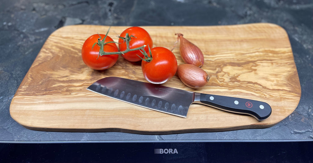 Naturform-Schneidebrett aus Olivenholz mit Tomaten, Schalotten und einem Küchenmesser auf dunkler Arbeitsfläche – stilvoll angerichtete Kochvorbereitung auf einem hochwertigen Brett.