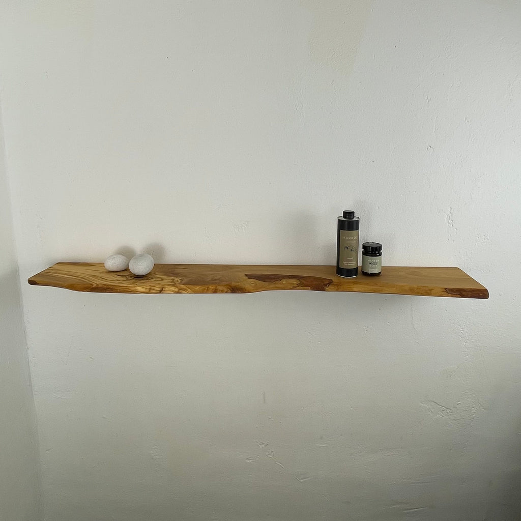 Massives Wandboard aus Olivenholz mit lebendiger Maserung, Naturkante und typischer Unregelmäßigkeit – montiert an einer weißen Wand, dekoriert mit MARKOS OLIVE Produkten.