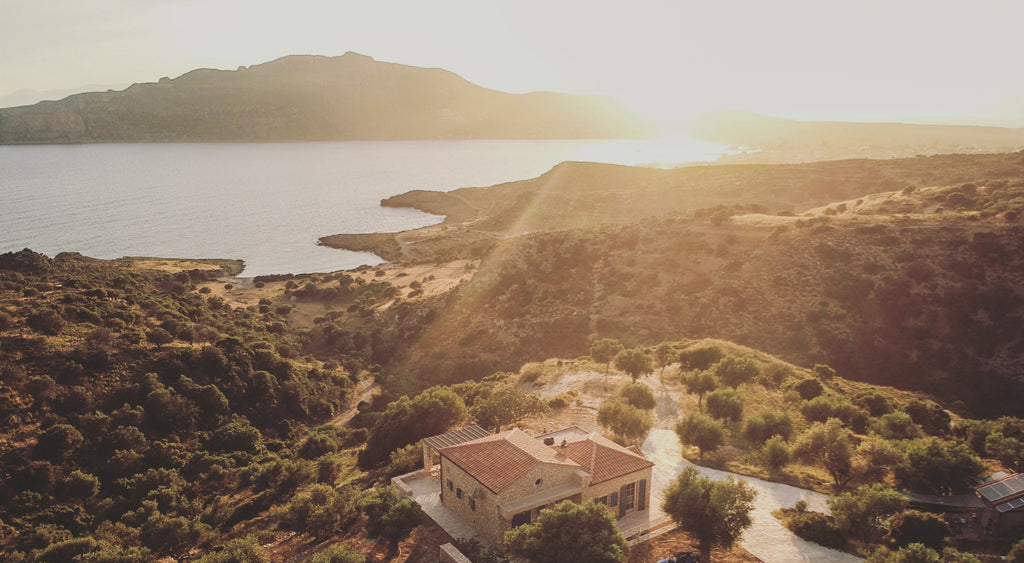 Drohnenaufnahme eines traditionellen Steinhauses inmitten eines Olivenhains an der Küste von Plitra, Griechenland. Die warme Abendsonne taucht die hügelige Landschaft und das Meer in goldenes Licht – ein stimmungsvoller Blick auf die Ursprungsregion von MARKOS OLIVE.