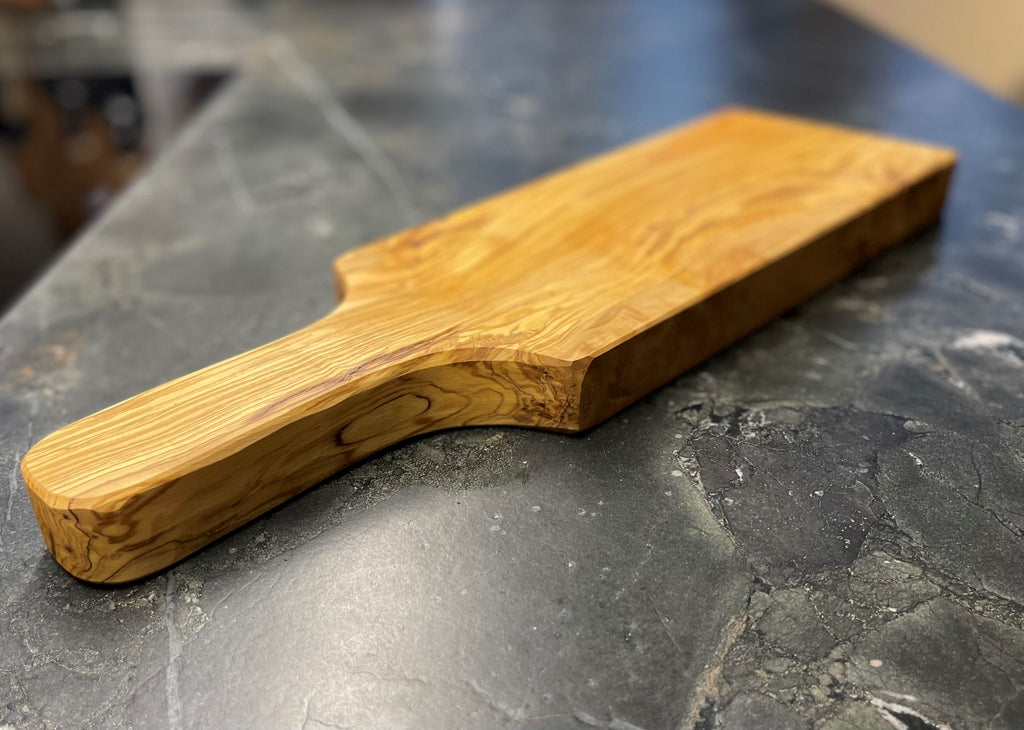 Seitliche Ansicht eines handgefertigten Schneidebretts aus massivem Olivenholz mit ergonomischem Griff, sanft abgerundeten Kanten und markanter Maserung – präsentiert auf dunklem Naturstein.
