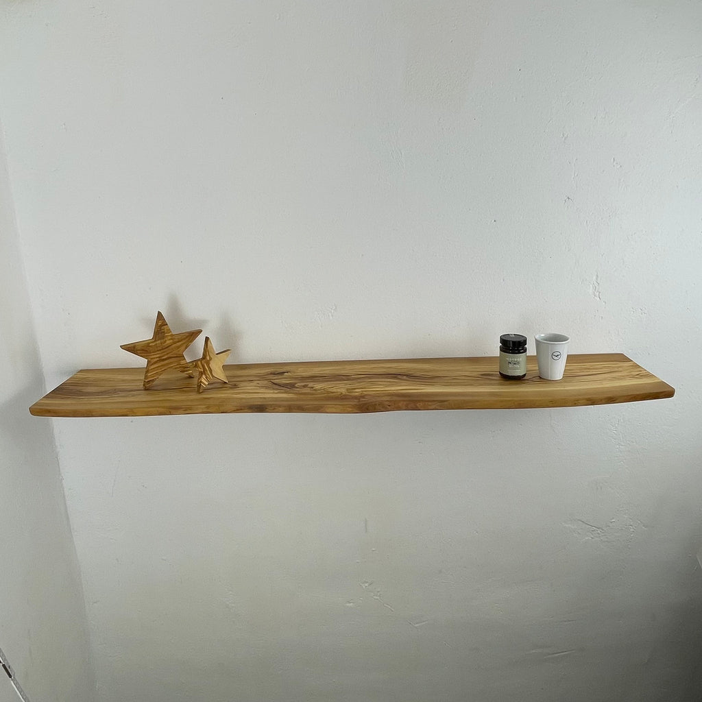 Langes Regalbrett aus Olivenholz mit sanfter Naturkante und markanter Maserung, dekoriert mit Sternen und kleinen Gefäßen – montiert an weißer Wand.