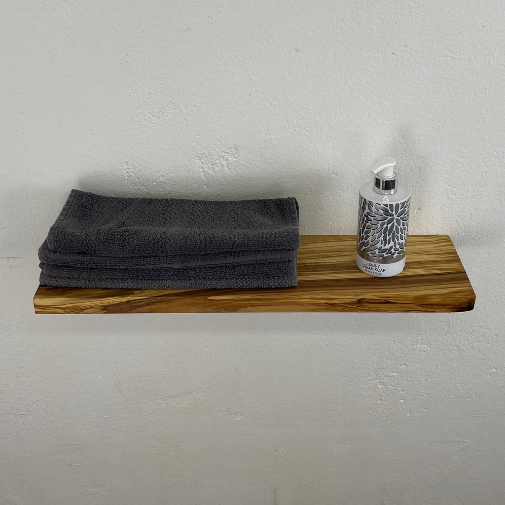 Geradliniges Regalbrett aus Olivenholz mit glatten Kanten und ruhiger Maserung, dekoriert mit grauen Handtüchern und Seifenspender – an weißer Wand montiert.